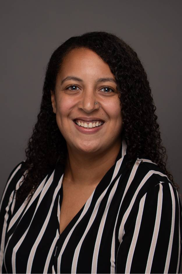 Justina Belle smiling for her GVSU headshot against with a dark gray background, wearing a black and white vertically striped blouse